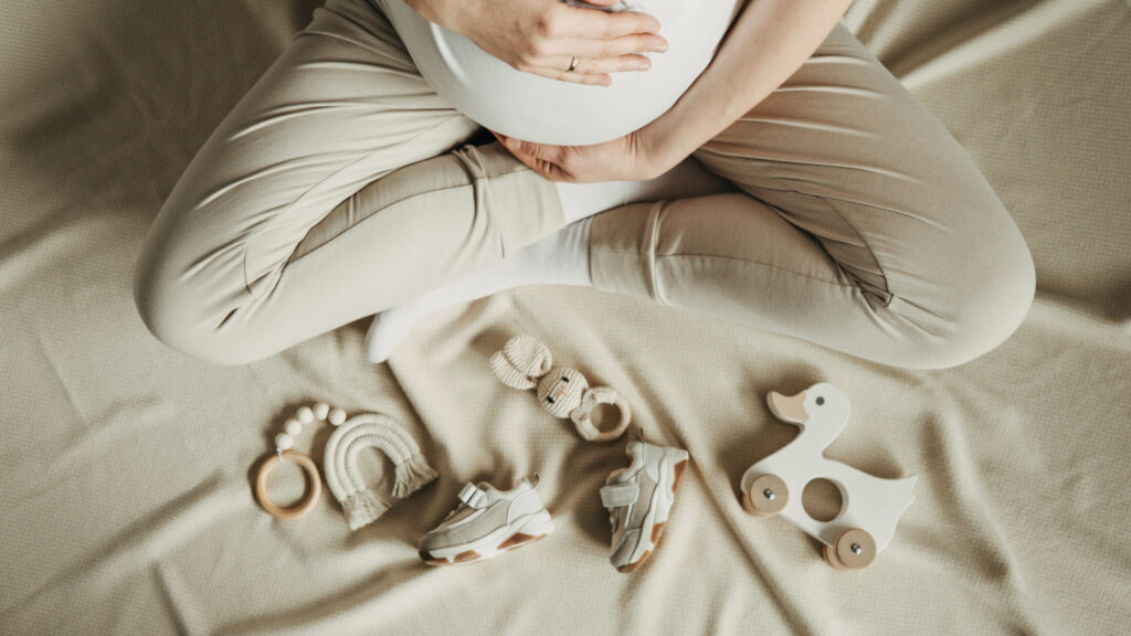 Έγκυος γυναίκα χαϊδεύει την κοιλιά της μπροστά από ξύλινα παιδικά παιχνίδια και παπούτσια
